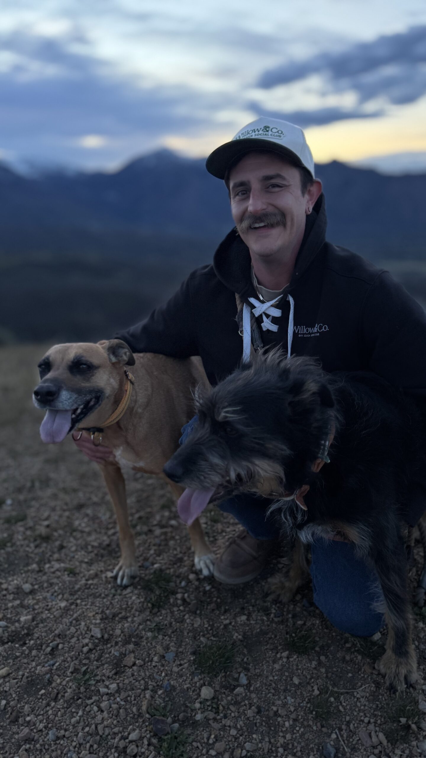 Tyler, founder of Willow & Co. Off Leash Social Club, crouches outdoors at dusk wearing a Willow & Co. hat and hoodie, smiling with his two dogs Willow and Cooper against a mountain backdrop in Salt Lake City, Utah. They are on the Deer Ridge Off Leash Trail in Draper